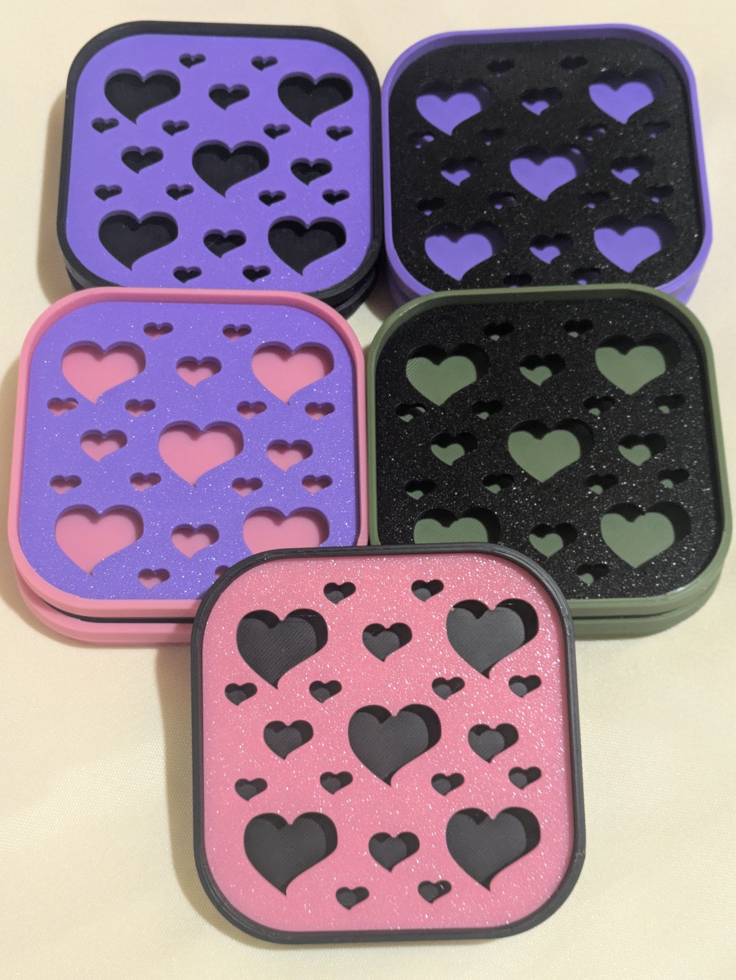 Five square 3D-printed soap trays with heart patterns in purple, pink, black, and green, arranged on a light beige background.