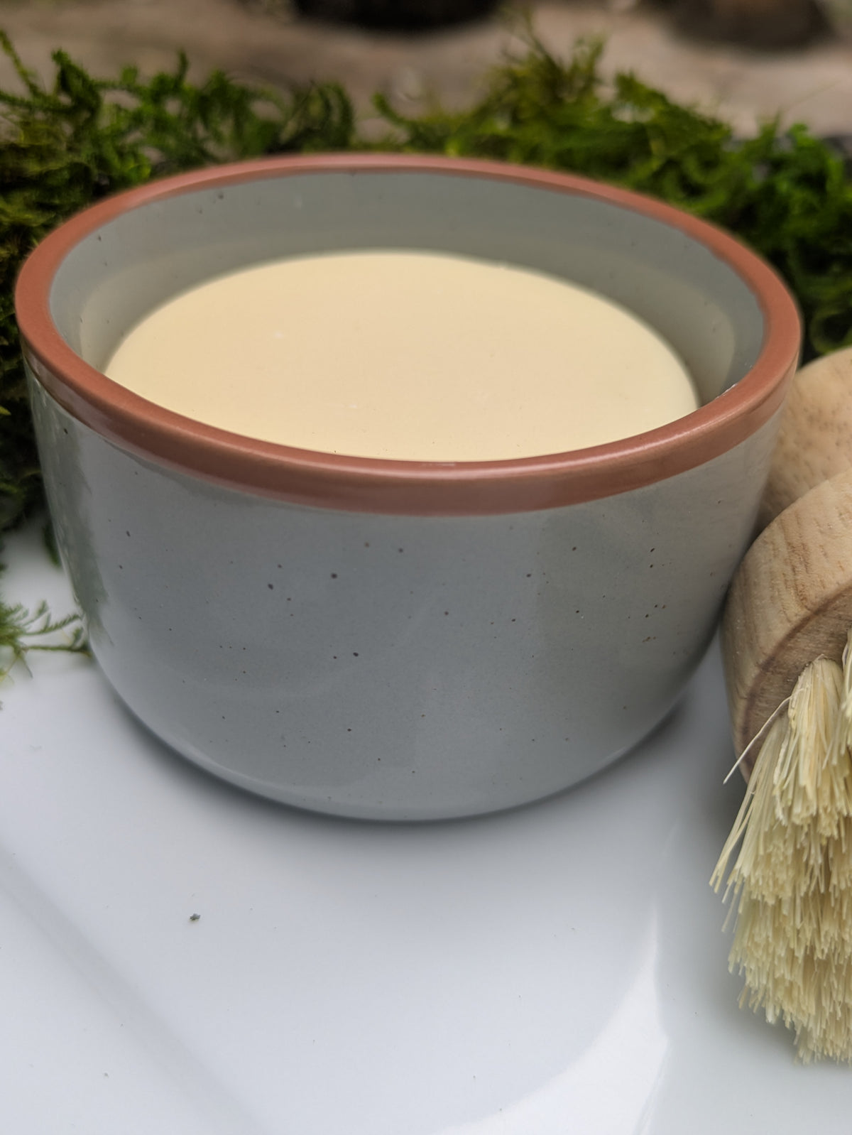 A small speckled blue ceramic bowl filled with a pale creamy soap, placed on a white surface with a wooden-handled brush in the foreground and green moss in the background.