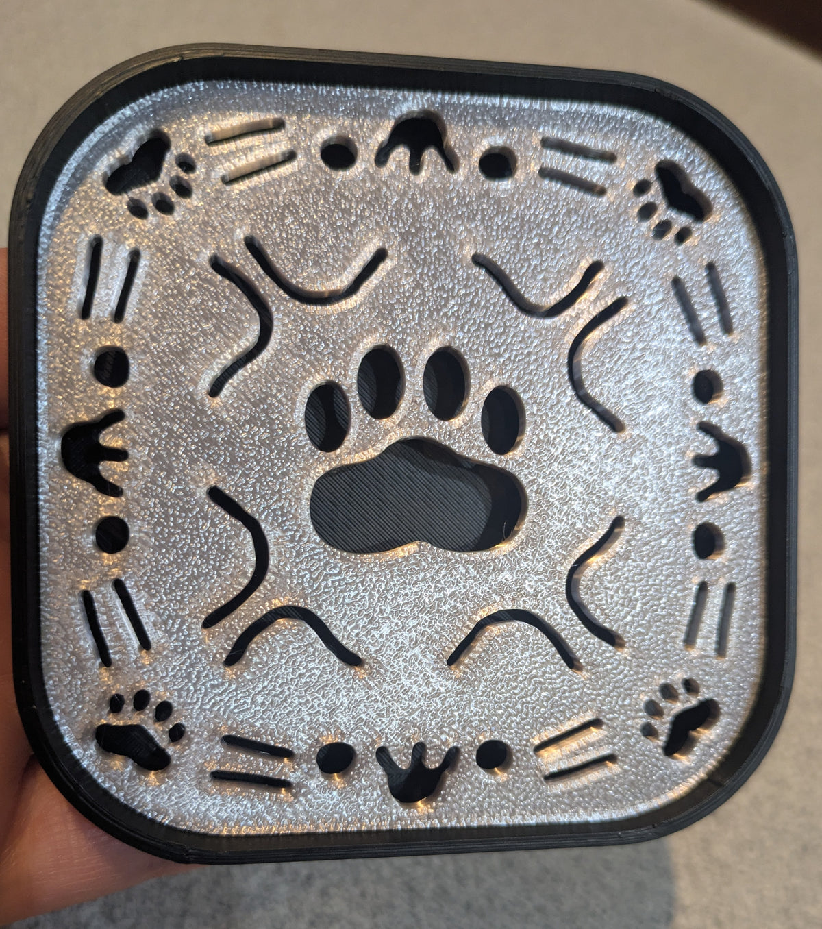 A square 3D-printed soap tray with rounded corners, featuring a gray/silver top with a black bottom contrast. Paw print in the middle, cat face cutouts on each side, along with small paw prints in the corners.