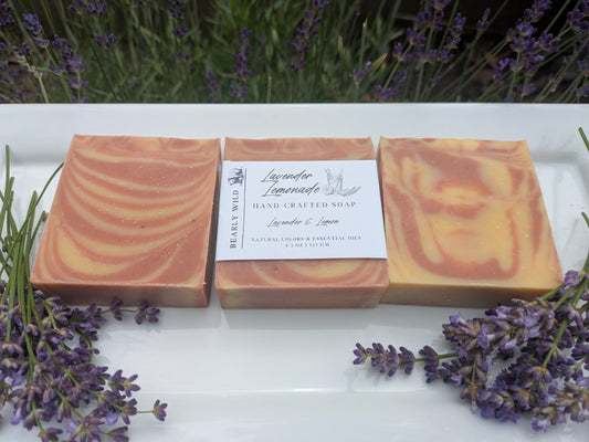 Three bars of soap with a lavender lemon scent on a white surface with lavender flowers. Soap has pink and yellowish-orange colors throughout, in different line formations.