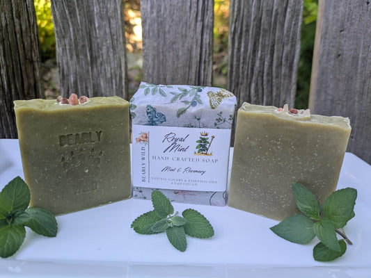 Three bars of handcrafted soap in a muted sage green color are displayed on a white surface, with of them being a wrapped bar. The soap bar have pink salt in a half circle resembling a crown and fresh mint leaves are placed in front.