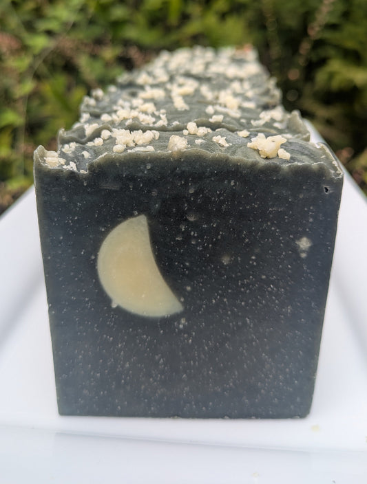 Soap on a white plate with a nature background. The bars resemble the night sky. Each bar is dark black and gray, white specks throughout to look like stars with a cream colored crescent moon.