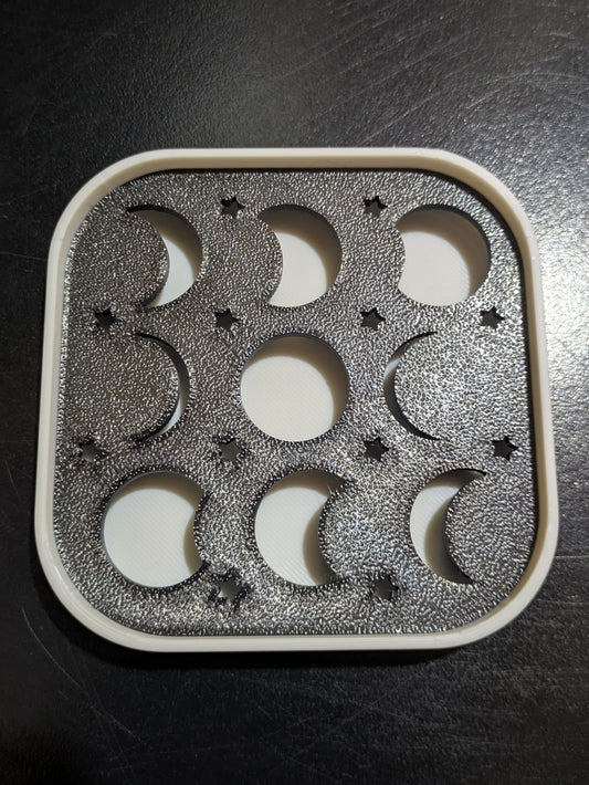 A square white plastic mold tray with a black insert featuring moon phases and star cutouts, photographed on a dark surface.