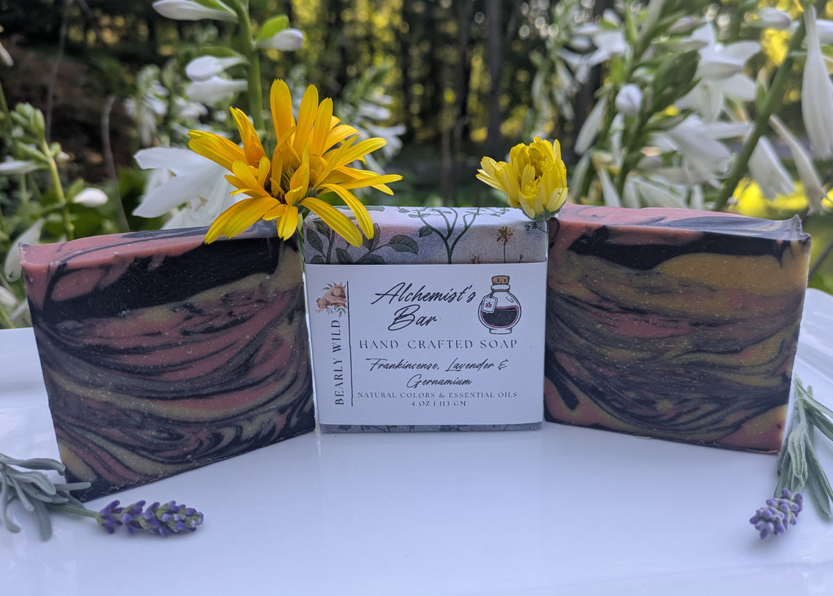 3 square shaped soaps with black, pink and yellow swirls throughout. The middle soap features a product label. Soaps are on a white tray and nature background. 