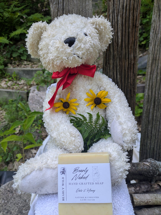 A cream-colored teddy bear plush with a red ribbon, plush covered with yellow flowers and ferns, holding a bar of soap labeled 'Bearly Naked' outdoors in front of a wooden fence and greenery.