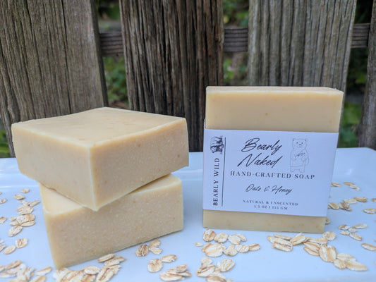 Two stacked bars of creamy beige handmade soap next to a wrapped bar of the same soap, labeled 'Bearly Naked Hand-Crafted Soap' and 'Oats & Honey,' displayed on a white surface scattered with oats, with a rustic wooden fence background.