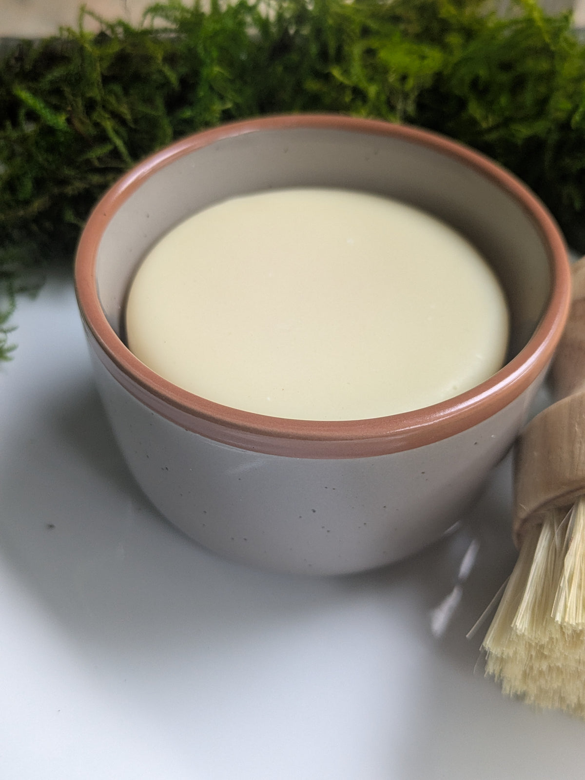 A small speckled beige ceramic bowl filled with a pale creamy soap, placed on a white surface with a wooden-handled brush in the foreground and green moss in the background.
