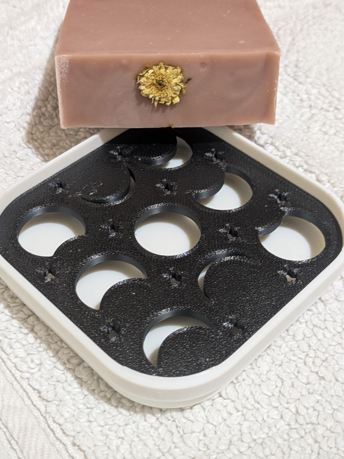A bar of Tranquil Mist which is a light pink soap with a small dried flower embedded in the top surface, resting on a white and black 3D-printed phases of the moon tray over a textured white towel.