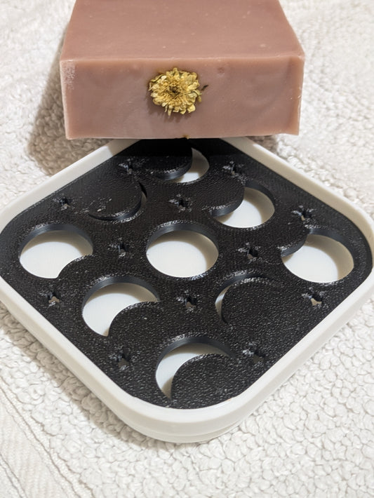 A bar of Tranquil Mist which is a light pink soap with a small dried flower embedded in the top surface, resting on a white and black 3D-printed phases of the moon tray over a textured white towel.