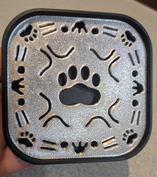 A square 3D-printed soap tray with rounded corners, featuring a gray/silver top with a black bottom contrast. Paw print in the middle, cat face cutouts on each side, along with small paw prints in the corners. 