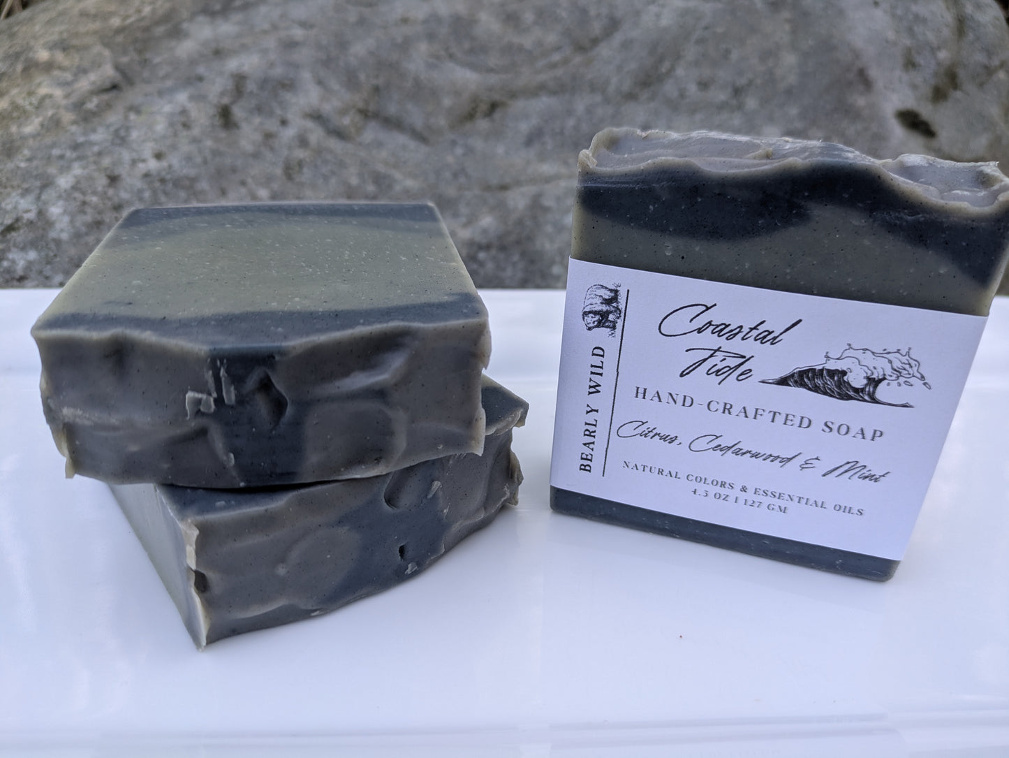 Three bars of grayish blue soap, 1 with a 'Coastal Tide' label on a white surface with a large rock in the background.