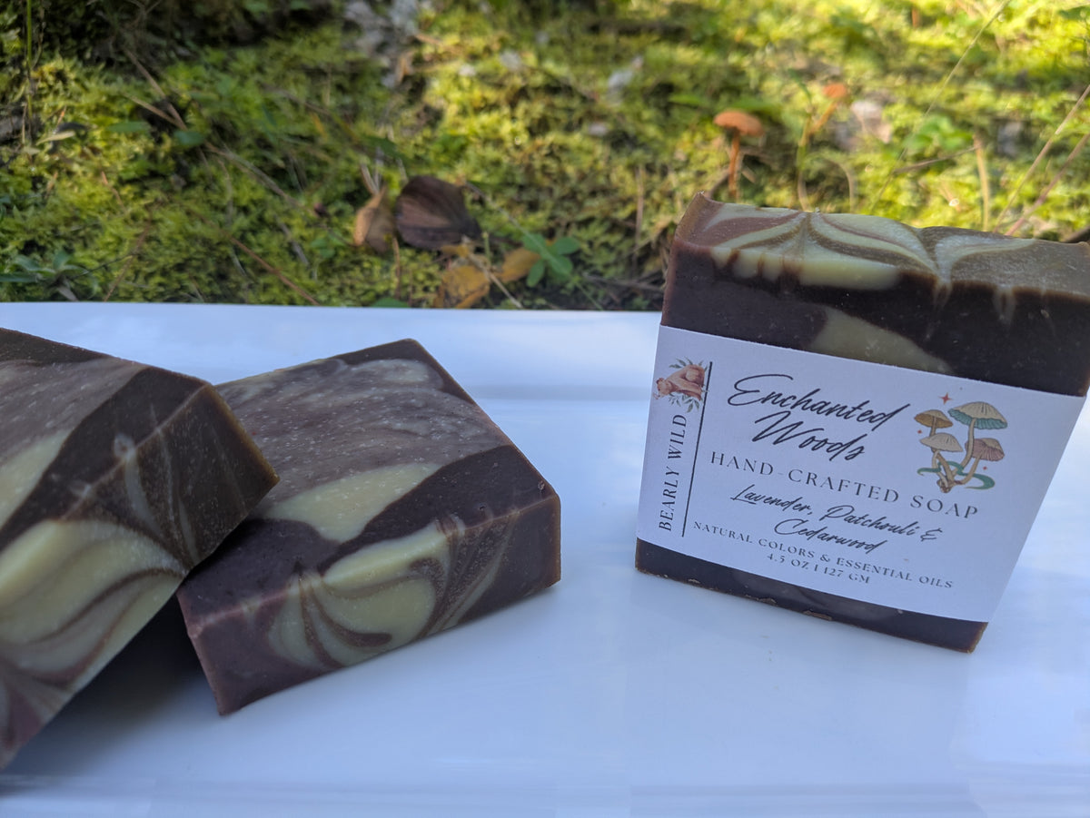 3 soap bars with purple, brown and cream swirls. Soap is on a white plate with nature in the background. 1 soap is labeled Enchanted Woods while the other 2 show the design. 