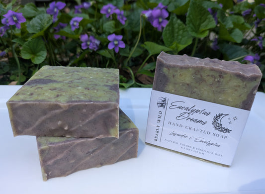 Three handcrafted soap bars in green and purple, 1 labeled on a white surface, surrounded by Violets. 