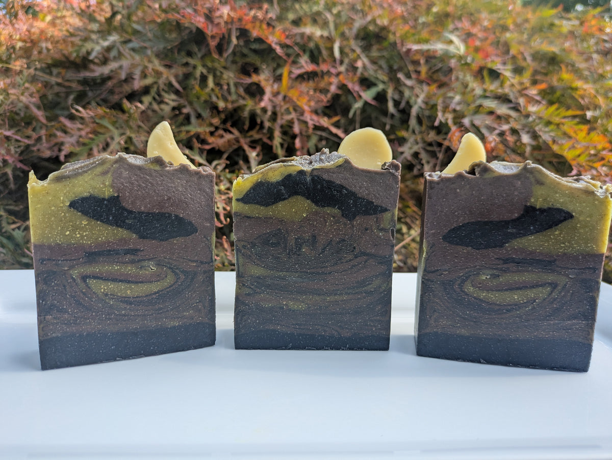 3 soaps on a white plate with a nature background . Swirled with black, dark purple and green. Two of the are topped with crescent moon and one with a full moon. 