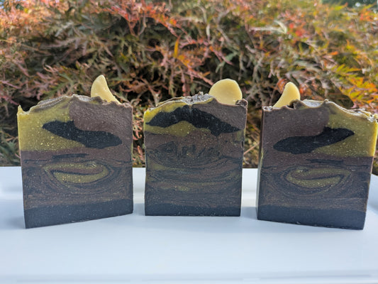 3 soaps on a white plate with a nature background . Swirled with black, dark purple and green. Two of the are topped with crescent moon and one with a full moon. 