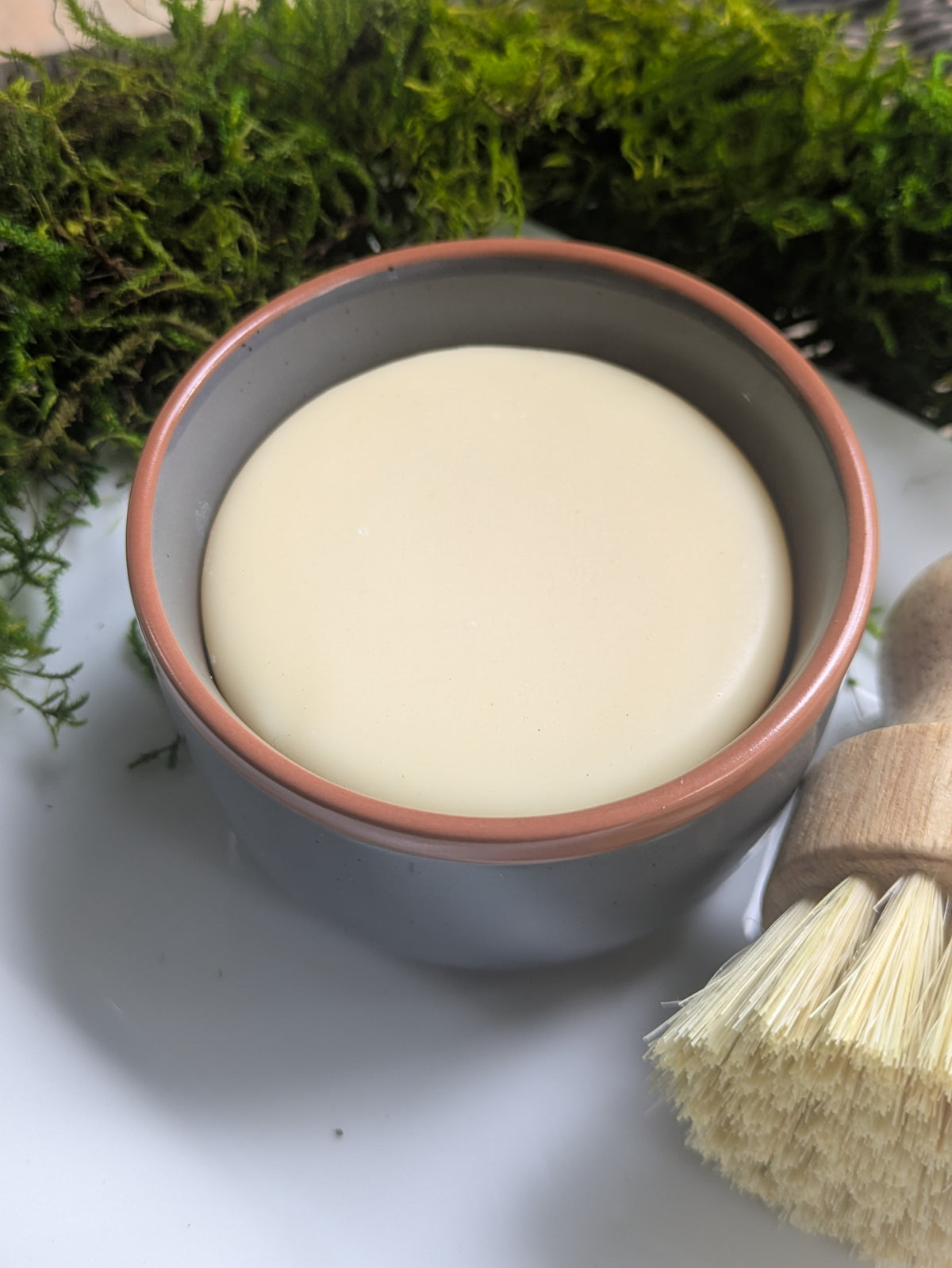 A small speckled dark gray ceramic bowl filled with a pale creamy soap, placed on a white surface with a wooden-handled brush in the foreground and green moss in the background.