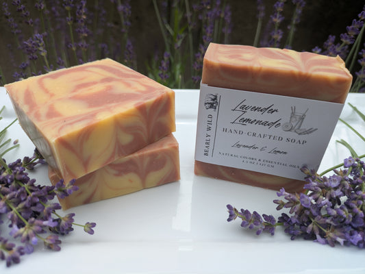 Three bars of Lavender Lemonade soap, surrounded by lavender plants in the background and lavender flowers on a white surface.