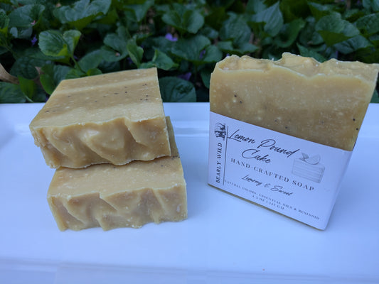 Three bars of yellow handmade soap, with one of them labeled "Lemon Pound Cake," on a white surface, with greenery in the background.