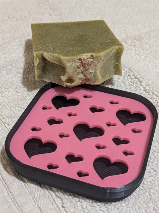 A rectangular bar of handmade soap in an olive green shade with pink salt on top, resting on top of a pink and black 3D-printed soap tray with heart-shaped cutouts, set against a textured white towel. 