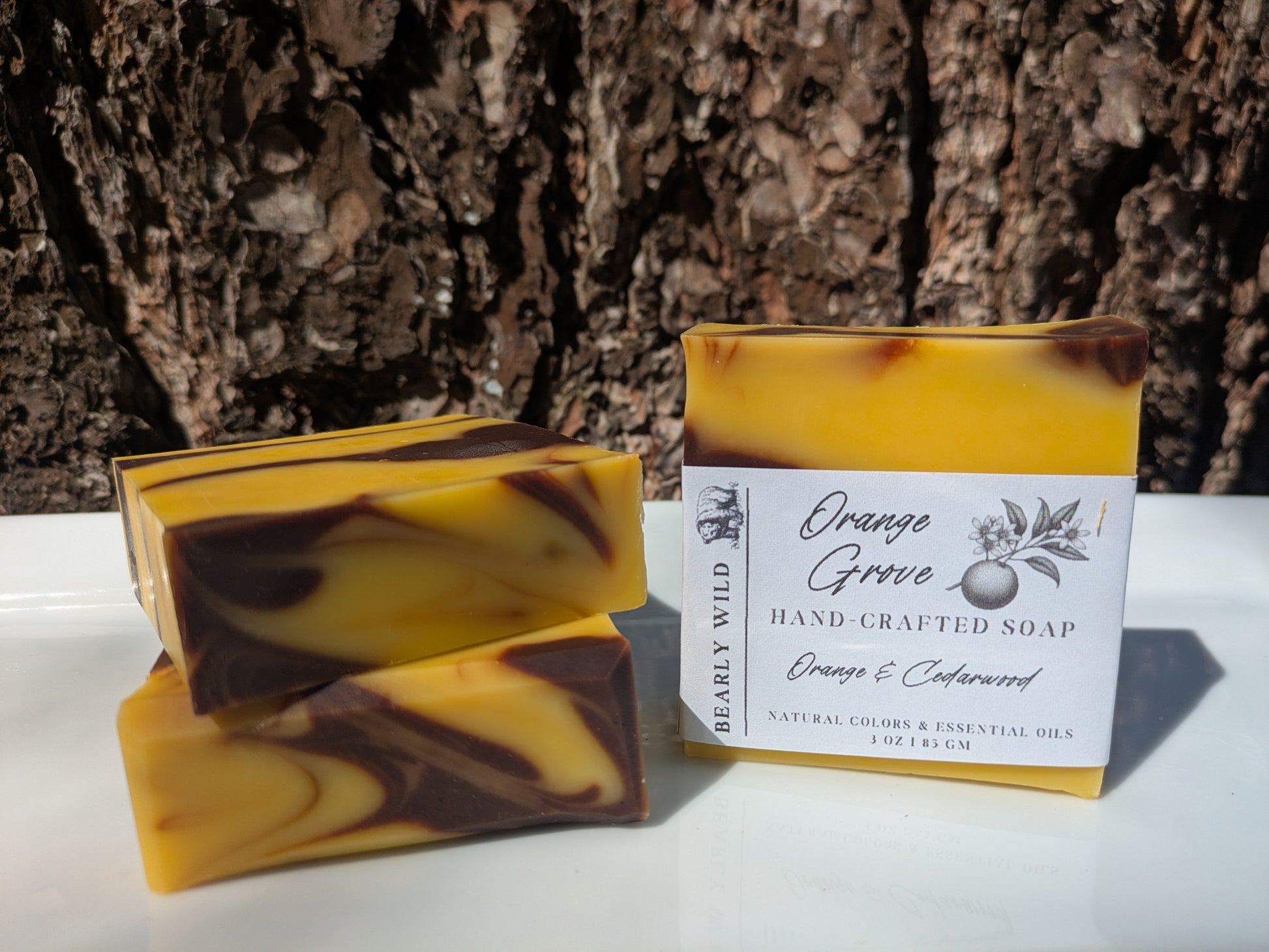 Three handcrafted soap bars with orange and brown lines and one with a brand label of Orange Grove, set on a white plate and against tree bark in the background.