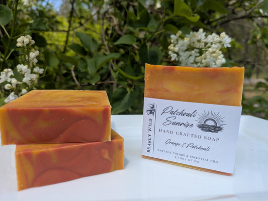 Three bars of orange, pink, and yellow swirled soap, 1 of them with a 'Tranquil Mist' label on a white surface with greenery in the background and white lilac flowers. 