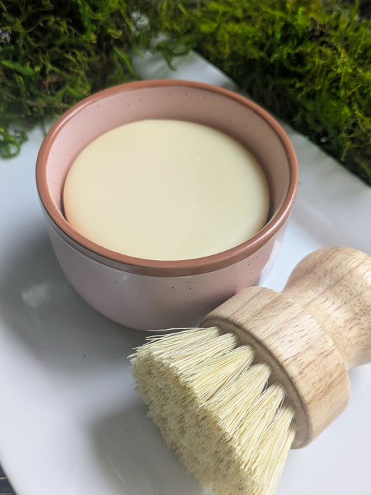 A small speckled pink ceramic bowl filled with a pale creamy soap, placed on a white surface with a wooden-handled brush in the foreground and green moss in the background.