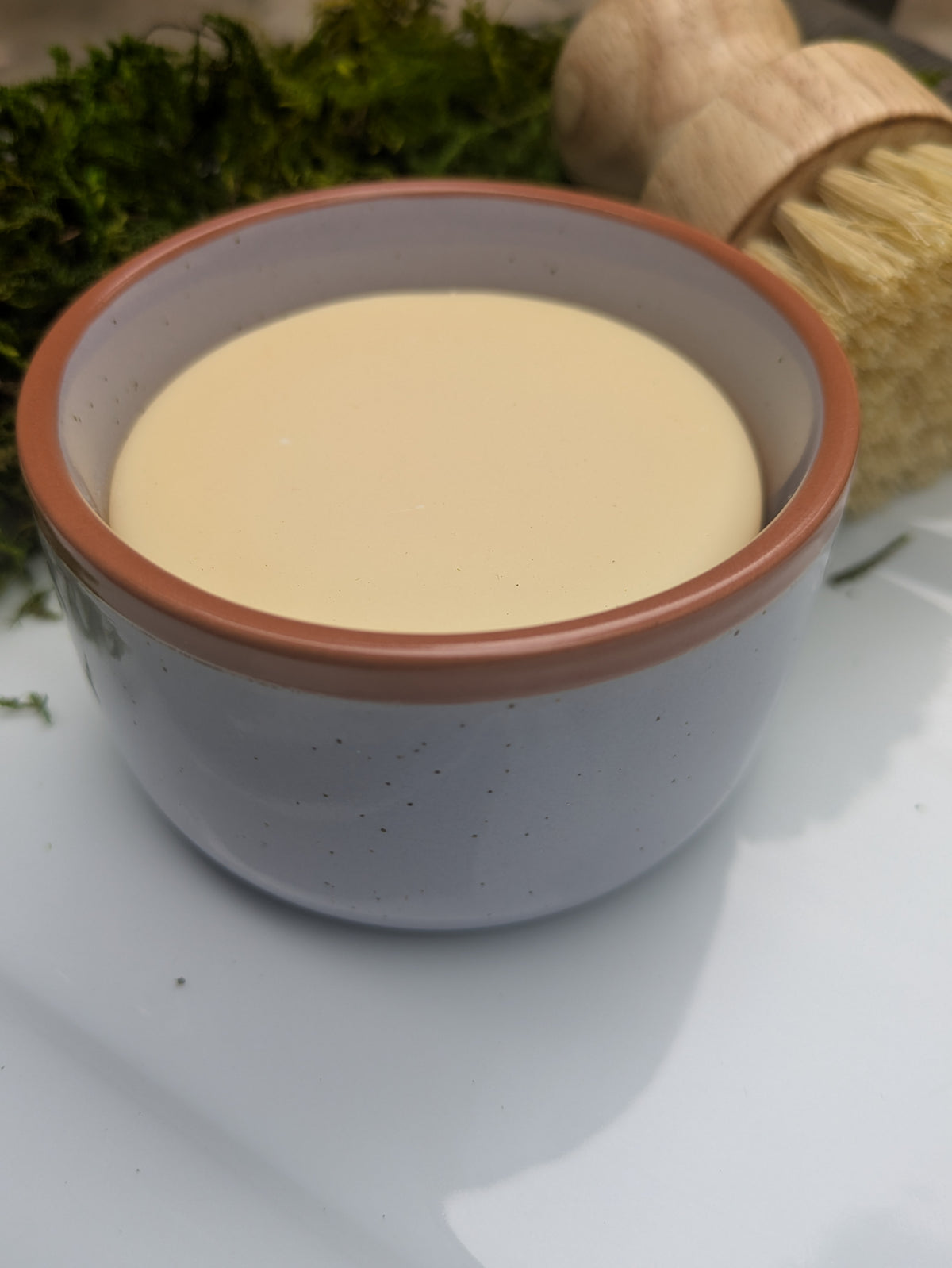 A small speckled purple ceramic bowl filled with a pale creamy soap, placed on a white surface with a wooden-handled brush in the foreground and green moss in the background.