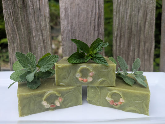 Three bars of handmade green soap with a half circle of pink salt to resemble a crown. They are stacked on a white surface, each topped with fresh mint leaves, with a blurred wooden fence and greenery in the background. 