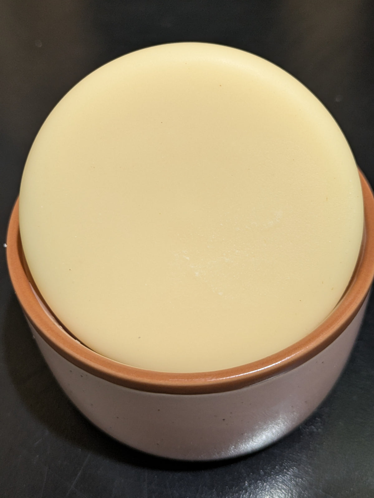 A round, solid bar of pale cream-colored dish soap in a speckled ceramic container,  set against a dark background.