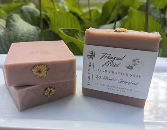 Three bars of pink soap with a white flower on top and 1 with a 'Tranquil Mist' label on a white surface with greenery in the background.