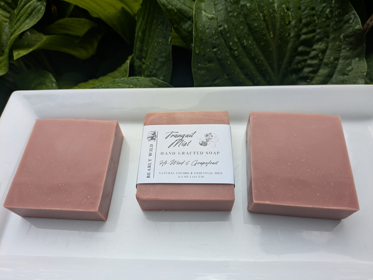 Three bars of pink soap with the middle bar featuring a 'Tranquil Mist' label on a white surface with greenery in the background.