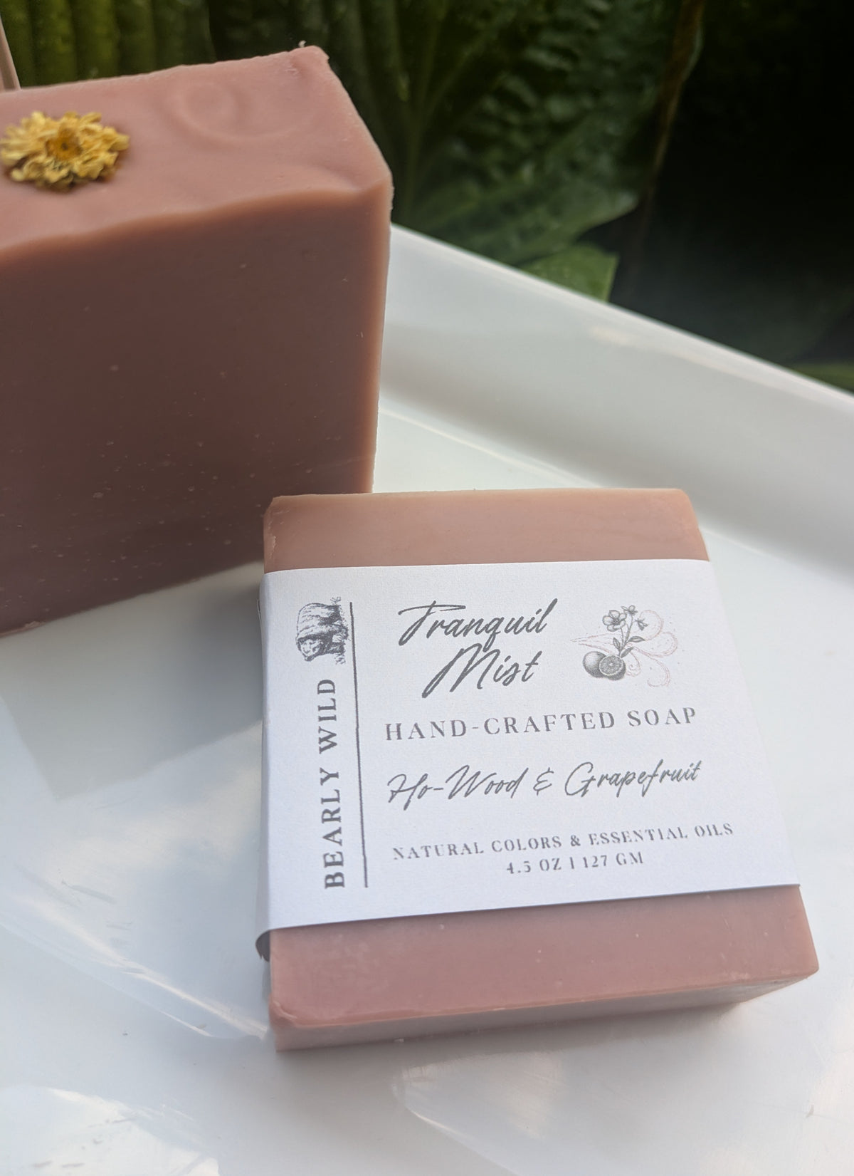 Two bars of pink soap with a white flower on top and 1 with a 'Tranquil Mist' label on a white surface with greenery in the background.