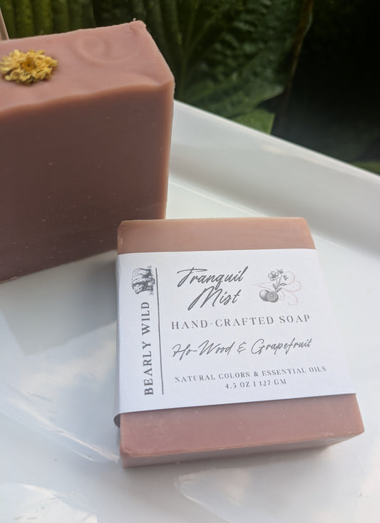 Two bars of pink soap with a white flower on top and 1 with a 'Tranquil Mist' label on a white surface with greenery in the background.