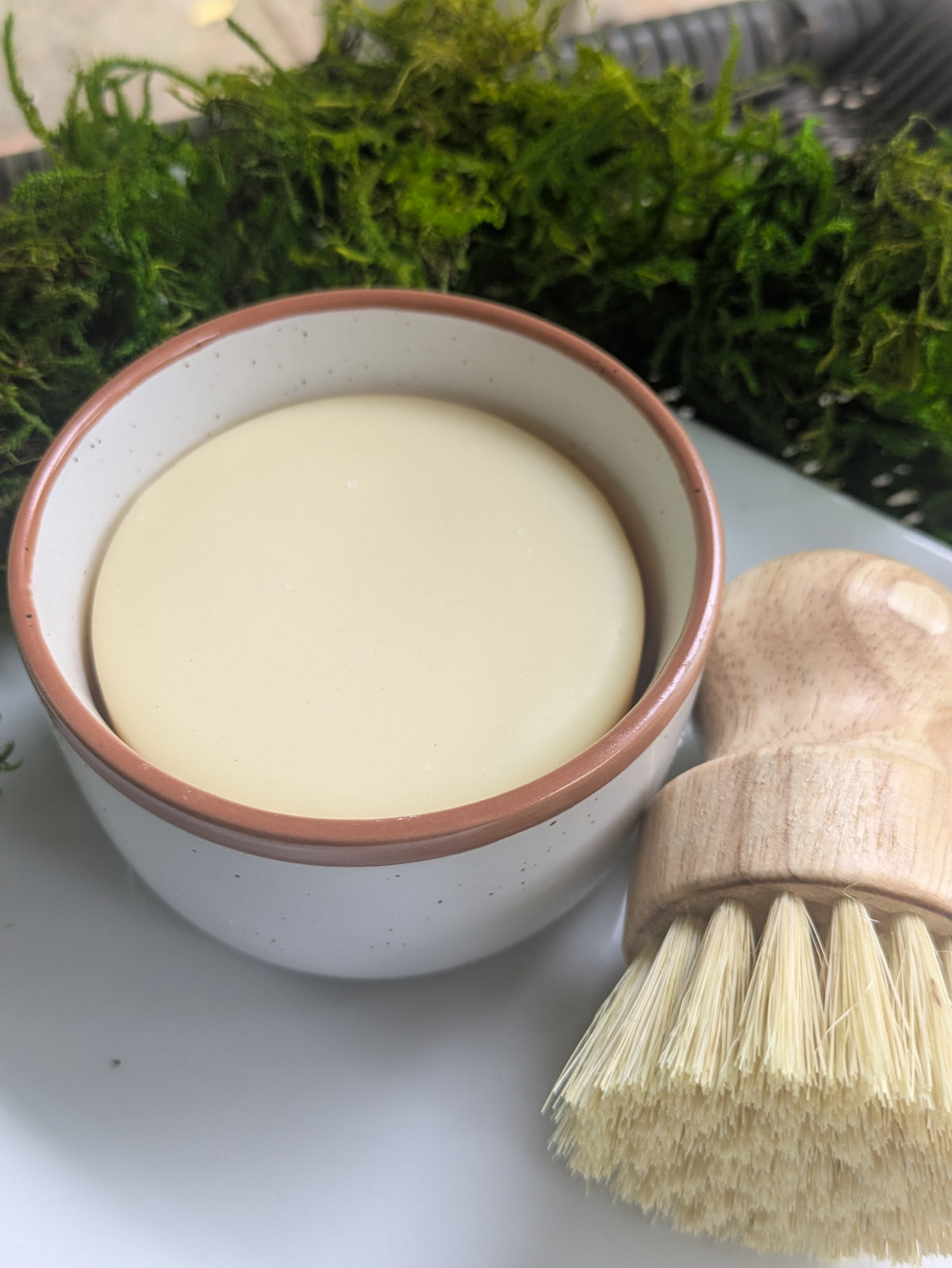 A small speckled white ceramic bowl filled with a pale creamy soap, placed on a white surface with a wooden-handled brush in the foreground and green moss in the background.