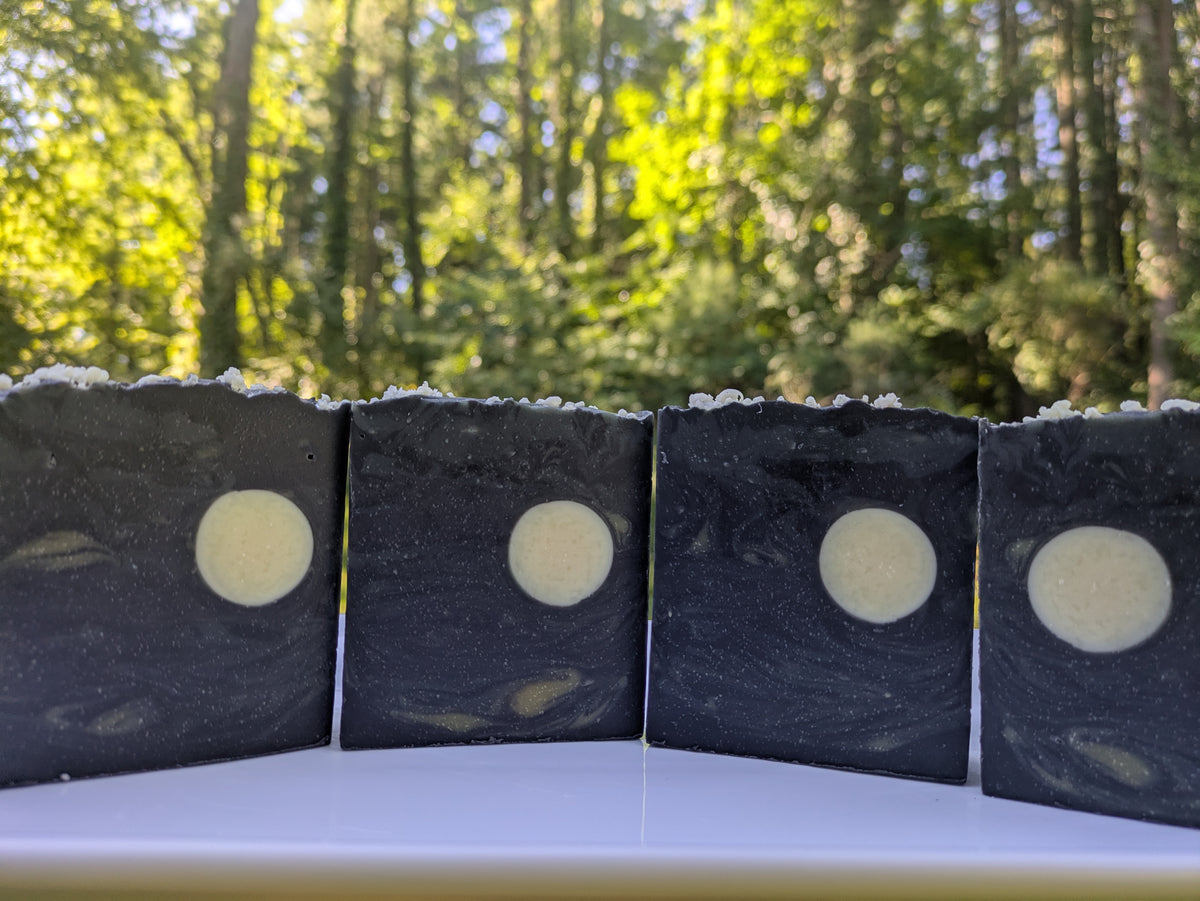4 bars of soap on a white plate with a nature background. The bars resemble the night sky. Each bar is dark black, gray and cream swirls with a full cream colored moon.  