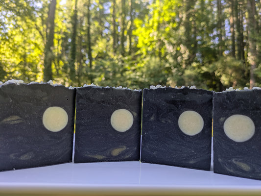 4 bars of soap on a white plate with a nature background. The bars resemble the night sky. Each bar is dark black, gray and cream swirls with a full cream colored moon.  